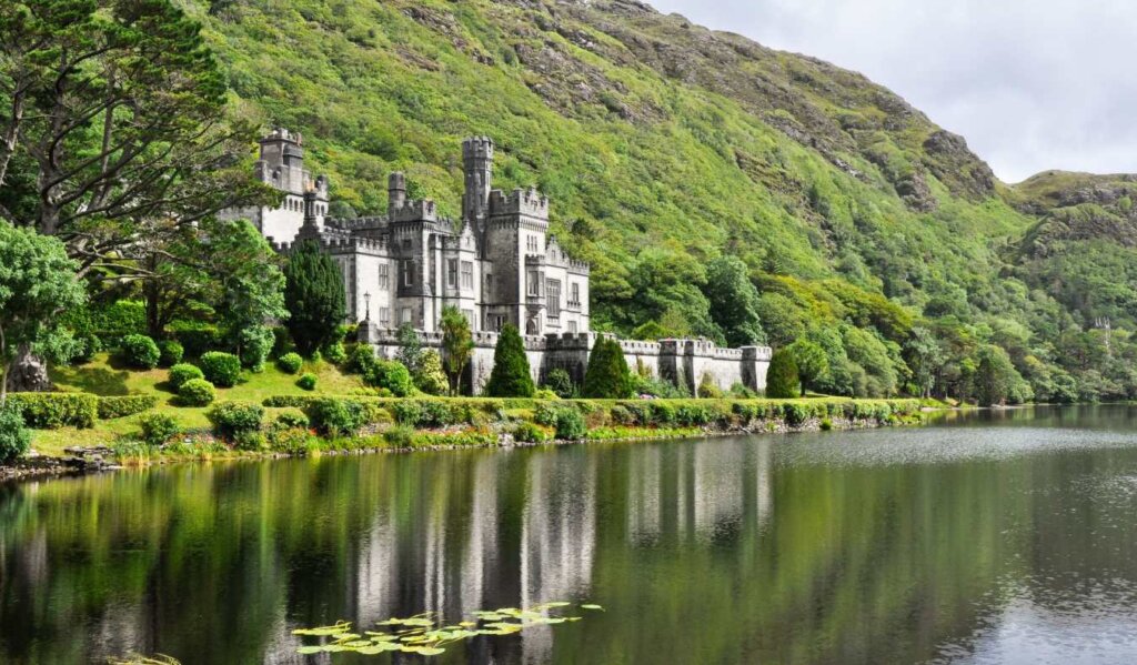 The castle-like Kylemore Abbey set against green hills, with a still lake in the foreground containing a reflection of the building